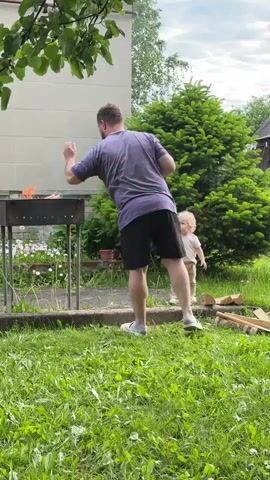 An adult male and a young child are situated outdoors in a grassy area, likely a backyard or garden. The man, seen from behind wearing a striped t-shirt, dark shorts, and light-colored shoes, is attending to a barbecue grill (brazier) on the left side of the frame. Smoke or steam rises from the grill, indicating a fire is burning. He briefly bends over the grill before standing upright again.

To the man's right, a young child with light, curly hair, dressed in a light-colored t-shirt and trousers, stands watching. The child remains relatively still throughout the video. The ground is covered in green grass, and several pieces of wood or logs are scattered on the right side of the frame.

In the background, a light-colored building with square paneling is visible behind the grill. To the right, a large, lush evergreen tree dominates the middle ground. Further back on the right, a darker building, possibly a shed or a small house, is visible with red fabric (likely laundry) hanging on a line. The sky is overcast, suggesting a cloudy day, possibly late afternoon or early evening. The scene depicts a calm, domestic outdoor activity in Красный Маяк, Russia.