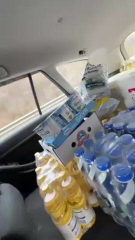 The camera is inside a car, turned around to face the back seat, which is filled with supplies. There are several packs of water bottles with blue caps, bottles of cooking oil with yellow liquid, a box of Skyr yogurt cups, and bags of food or supplies. The light outside is dim, suggesting an overcast day.
