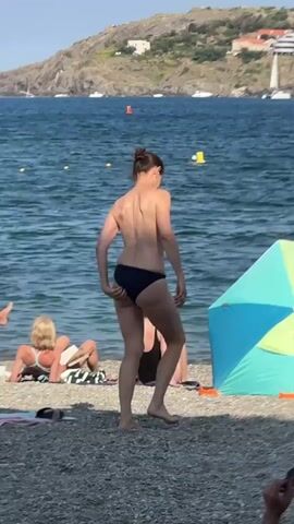 This media file captures a sunny day at a beach in Collioure, France. The scene is bustling with activity, indicating it's likely the middle of the day under clear skies. The water is a deep blue, with several boats, including a sailboat, visible in the distance, suggesting a calm sea. The shoreline is pebbly, characteristic of many Mediterranean beaches.

The main focus is a woman in a dark blue bikini walking across the beach. Her back is to the camera, and she appears to be moving away from the water. To her left, a woman in a black swimsuit is seated on the pebbles, reading a book. Further left, another woman with blonde hair is also seated, facing away from the camera, with a towel and possibly a book beside her.

On the right side of the frame, a colorful beach umbrella, predominantly blue and green with yellow accents, provides shade. A man in black swimming trunks stands near the umbrella, holding what appear to be sunglasses. In the background, a group of people are gathered on a small jetty or platform in the water, some appearing to be children. Another person is swimming further out in the sea, marked by a green ring.

The backdrop features a hillside dotted with buildings, typical of coastal towns in the region, with the distinctive French architecture. The overall atmosphere is relaxed and summery, with people enjoying a day by the sea. There is no visible text in the media file.