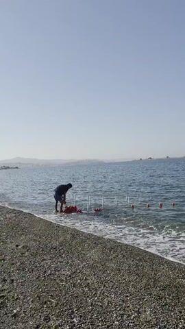 A person's shadow is visible on a pebble beach as gentle waves lap the shore. In the background, a calm blue sea stretches towards a hazy horizon, dotted with small buoys marking a swimming area. The coastline is lined with buildings, suggesting a populated area. The sky is clear and bright, indicating a sunny day.