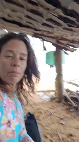 A woman with shoulder length brown hair speaks toward the camera from under the dilapidated roof of a beachside structure. The roof is constructed of corrugated metal and supported by wooden posts. The ground is sandy and strewn with debris including sticks and palm fronds. The camera pans up to show the structure's roof, then the ocean visible beyond. The ocean is choppy with white waves. The day appears overcast.