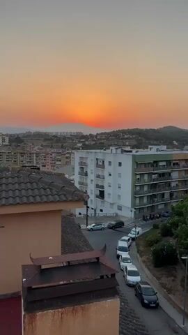 A vertical video shows a sunset over a residential area. The sun is a bright orange disc behind distant mountains. In the foreground, a tiled roof and a chimney are visible. Below the roof, a street curves through a neighborhood of apartment buildings. Several cars are parked along the street or driving past. The sky is a gradient of orange, pink, and pale yellow. The buildings are a mix of white, beige, and green, with balconies visible on some. Trees and greenery are interspersed throughout the scene. The overall atmosphere is peaceful and serene.