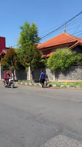 A woman rides a motorcycle down a paved street on a sunny day. She is wearing a red jacket and brown pants. Behind her, a man in a blue shirt stands near a stone wall. Trees and houses with orange roofs are visible in the background.