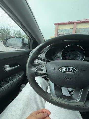 A POV shot from the driver's seat of a Kia car, with the driver's legs visible in light gray sweatpants. The steering wheel and dashboard are in focus. Outside the window, it is raining, and a parking lot is visible, along with a building in the background. The car is parked.