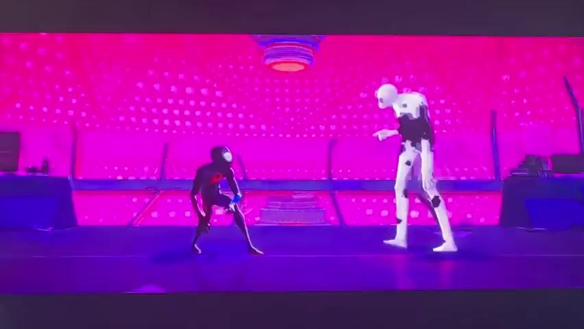 The video appears to be a scene from an animated film, likely "Spider-Man: Into the Spider-Verse" or a similar franchise, given the distinctive art style. The primary subjects are two characters resembling Spider-Man. On the left is a character in a black and red Spider-Man suit, and on the right is a character with a white body and black splotches, resembling the Spot.

The scene is set in a vibrant, abstract environment characterized by intense pink and purple lighting, creating a surreal and energetic atmosphere. The background features a textured pattern of small, circular or polygonal shapes, adding to the otherworldly feel. In the center of the frame, there's a tiered, cake-like object, its purpose unclear within this context. Above this, a circular structure with radiating elements hangs from the ceiling.

The activity depicted is a confrontation or interaction between the two Spider-Man characters. The character on the right, the Spot, appears to be in motion, with his splotches seemingly detaching or moving dynamically. The character on the left is posed defensively or observantly. The overall mood is intense and action-oriented, with the swirling, colorful tunnel at the end suggesting travel through different dimensions or realities, a common theme in Spider-Man multiversal stories. The visuals suggest a moment of high stakes and visual spectacle. There is no visible text within the frames provided. The time of day and weather are not discernible due to the stylized and abstract nature of the setting.