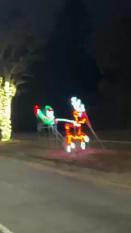 The video depicts a roadside scene at night, adorned with Christmas lights. A blurred image of a lawn decoration featuring an elf holding reins connected to a reindeer is visible, both illuminated with colorful lights. Further down the road, a row of trees are wrapped in bright, warm-toned lights. The road appears to be paved and is darker in color. The overall atmosphere is festive due to the holiday decorations.