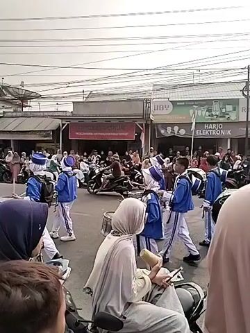 The video shows a street scene with a marching band, likely a drum corps, parading. The band members are wearing matching blue and white uniforms with hats. They are carrying various drums, including snare drums and bass drums, and appear to be playing them. Spectators are lining the street, watching the parade. Some spectators are seated on motorcycles, while others are standing. Buildings are visible on either side of the street, with shops and signage. There are power lines and utility poles overhead. The weather appears to be overcast. The video is shot from a slightly elevated perspective, looking down at the parade. The marching band is moving from left to right across the frame. Some members of the band are playing instruments, while others are holding them. The spectators are looking towards the band and some are holding up phones to record the event. The scene is dynamic and filled with activity.