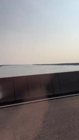 The video shows a view from a moving vehicle, likely a car, crossing a bridge over a wide river. The camera is positioned to capture the scene through a concrete barrier separating the road from the river. The water is calm and appears wide, with trees and vegetation visible on the distant banks. The sky is clear and bright, suggesting a sunny day. Traffic signs are visible on a lamppost, including a yellow diamond-shaped sign indicating a narrowing of the road ahead, with a supplementary rectangular sign below it. The perspective shifts slightly as the vehicle moves, offering glimpses of the river and its surroundings. The road surface in the foreground is asphalt. No people are visible in the frame. The video appears to be a short recording of a scenic drive.