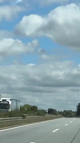 The video shows a highway with multiple trucks and cars driving on it. The sky is partly cloudy, with large white clouds against a blue sky. The highway has two lanes in each direction, separated by a concrete barrier. There are trees and bushes on either side of the highway. The camera is looking forward from a vehicle on the highway, so the road and surrounding scenery are moving past. The video appears to be a recording of a travel experience.