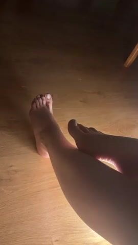 THE VIDEO SHOWS A PAIR OF BARE FEET ON A WOODEN FLOOR. THE FEET ARE SHOWN FROM A SLIGHTLY ABOVE AND TO THE SIDE ANGLE, WITH THE LIGHT CASTING A SHADOW FROM THE LEGS ONTO THE FLOOR. ONE FOOT HAS WHITE NAIL POLISH ON ITS TOES, WHILE THE OTHER HAS DARKER NAIL POLISH. THE TOES OF THE FEET ARE SLIGHTLY MOVED DURING THE VIDEO.