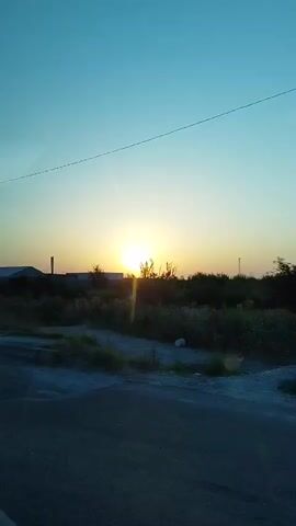 A time-lapse video shows a sunset over a semi-arid landscape. The sun, a bright orb, is partially obscured by low-lying vegetation and appears to be setting behind a distant structure. The sky is a gradient of blue and yellow, transitioning from a clear blue at the top of the frame to a warmer, yellowish hue near the horizon. In the foreground, a dark, paved surface, possibly a road, runs horizontally across the bottom of the frame. The surrounding area is covered in dry grass and sparse bushes, with a few scattered white objects visible. A thin wire stretches across the sky in the upper portion of the frame. The overall scene suggests an open, rural environment at dusk.
