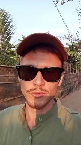 A man wearing sunglasses and a brown baseball cap is walking outside on a sunny day. He has a mustache and a goatee, and is wearing a green collared shirt. He is walking along a street with trees and a bamboo fence. He is looking directly at the camera and speaking.