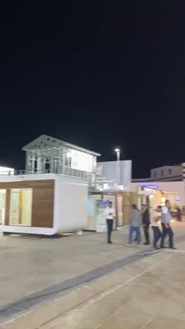 The video opens with a blurry, dark night scene. As the camera pans, it reveals an outdoor plaza or walkway. Several people are walking around, some in groups, others individually. There are buildings in the background, some appearing to be stalls or shops, with lights illuminating them. A tall pole with lights stands prominently in the foreground on the right side. A row of flags on poles is visible, with some flags displaying yellow and white designs, and others showing blue and white. One flag clearly reads "BUILD". The ground is paved with large tiles. The overall impression is of an outdoor event or gathering at night.