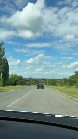 This is a vertical, first-person perspective video captured from inside a car. The car is driving on a paved road, seemingly in a rural area. The road has a dashed white line indicating lanes. Ahead, another car, a dark SUV, is also traveling in the same direction, further down the road. The surrounding landscape is green with trees lining both sides of the road and fields beyond. The sky is a bright blue with large, fluffy white clouds, suggesting it is daytime and the weather is fair. There are no people visible, and the focus is on the road and the drive. The scene evokes a sense of journey or travel. The location is identified as Shamraivka, Ukraine. No visible text is present in the video.