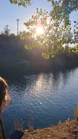 A video recording shows a body of water, likely a river or lake, with sunlight glinting off its surface. The sun is setting or rising, casting a warm glow through the leaves of a tree in the foreground. The tree is on a dirt bank overlooking the water. There are some small boats or kayaks on the water in the distance, and the opposite bank of the water appears to be lined with trees and some buildings. The video captures the serene beauty of the natural landscape as the day transitions.