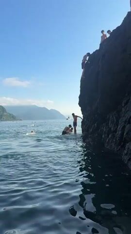 The video shows a group of young people enjoying a sunny day at a rocky coastline. The camera is positioned low, close to the water's surface, giving a sense of immersion. In the foreground, the blue water ripples gently, reflecting the bright sky. A large, dark, rocky cliff dominates the right side of the frame, with several individuals perched on its upper edge and midway down. One person is about to jump from the cliff into the water below.

In the middle ground, a few people are in the water, some swimming and others gathered near the base of the cliff. One person appears to be assisting another into the water from the rocks. Further out in the water, more people are visible, enjoying recreational activities.

The background features a serene seascape with distant, lush green mountains shrouded in a light mist under a clear blue sky with a few scattered clouds. A small boat is visible on the horizon. The overall atmosphere is one of summer leisure and adventure.