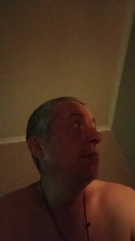 The video shows a close-up of a man's face. The man has graying hair and is looking upwards and to the right in the first frame. In subsequent frames, he turns his head towards the camera and makes a series of facial expressions, including a wide smile and a confused look. The lighting in the video is dim and casts shadows on the man's face. The background appears to be a wall or ceiling. The man is shirtless.