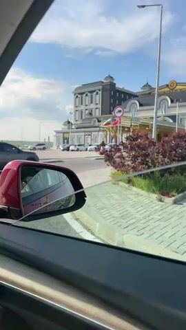 The video captures a view from inside a car of a luxurious building, likely a casino, with a large parking lot in front. The car is moving, and the building comes into full view as the vehicle proceeds. The building is ornate, with gold accents and multiple stories. There are several cars parked in the lot, and a few are seen driving on the road in front of the building. The sky is bright blue with a few white clouds, indicating a clear day. The car's side mirror is visible in the bottom left corner of the frame.