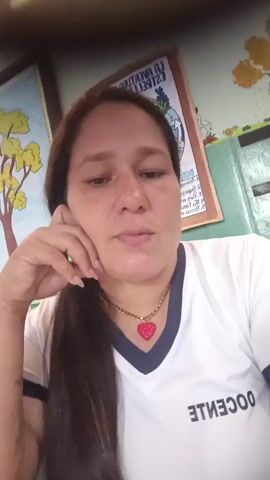 The video shows a close-up, selfie-style shot of a woman with long dark hair. She is wearing a white t-shirt with a dark blue collar and the word "DOCENTE" written in white lettering on the front. A gold chain with a pink heart-shaped pendant is visible around her neck. The woman's left hand is resting on her cheek, and she occasionally touches her face or lips with her fingers. In the background, there are two posters on a wall. One poster on the left depicts a tree with yellow leaves. The other poster, on the right, has text and an illustration of a bird. The environment appears to be indoors, possibly a classroom or office setting. The woman's facial expressions change throughout the short clip, transitioning from a neutral gaze to a smile and then back to a thoughtful expression.