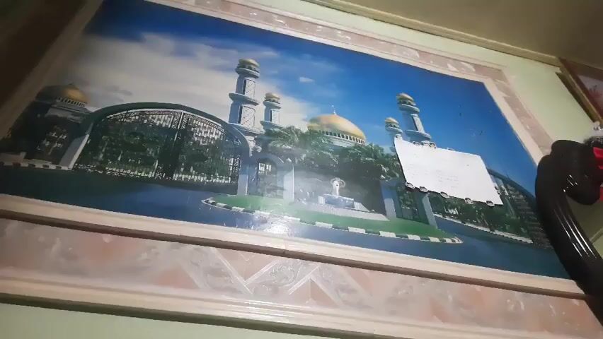 The video shows a framed picture hanging on a light-colored wall. The picture depicts a mosque with golden domes and minarets, set against a blue sky. In front of the mosque is a gated entrance, a fountain, and greenery. A white note is taped to the picture. To the right of the picture, part of a dark-colored object, possibly furniture, is visible. Sounds of a baby are heard.