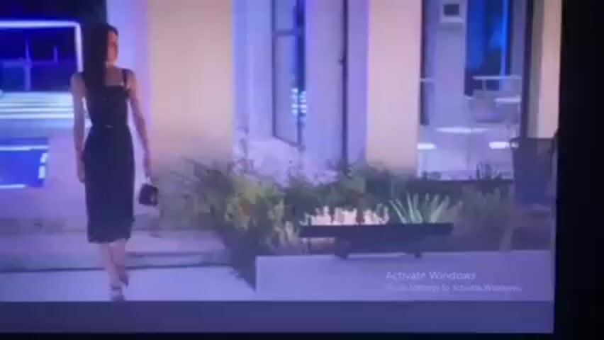 The video shows an outdoor setting at night, likely a courtyard or patio area adjacent to a building. There are three visible individuals. A woman in a black and white polka dot dress is walking toward the camera. In the background, there is a woman sitting at a table and a man sitting in a chair behind her.  There appears to be a swimming pool in the center of the courtyard. There is decorative lighting near the pool, and other decorative elements like plants and a fire pit. The overall color tone of the scene is cool, with blues and purples dominating the lighting. There's an "Activate Windows" watermark visible on the screen.