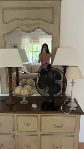 A woman in pink pajamas is standing in front of a mirror, taking a photo of herself with her phone. The mirror is mounted on a wooden dresser. On top of the dresser are two lamps with white shades, a decorative bowl filled with round objects, and a black electric fan. The reflection in the mirror shows a living room with a couch and a window. The woman raises one arm in the air while taking the photo.