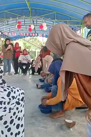 The video shows a group of people participating in an outdoor event. In the foreground, several women are crouched down, each with a blue bowl in front of them, and appear to be washing their feet or manipulating something with their hands in the water. One woman in a blue hijab and an orange tunic is particularly visible, actively engaged in this task. A halved coconut lies on the ground near her. Behind the participating women, other individuals are standing and observing, some holding up phones to record. The event takes place under a blue canopy decorated with red and white balloons and flags. The background suggests a rural or semi-rural setting with buildings and trees visible. The overall atmosphere seems festive and communal.