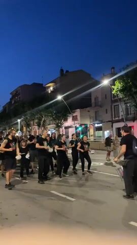 A group of approximately 15 people are walking down a street at dusk. They are all wearing black t-shirts with a colorful logo on the front and black pants or shorts. Most of them are holding drumsticks and appear to be playing drums or other percussion instruments. Some of them are also holding small tambourines. The group is moving in formation, with a leader at the front of the group who is also holding drumsticks and appears to be directing them. The street is paved and there are buildings on either side of the street. There are streetlights on, illuminating the scene. Some of the buildings have windows and balconies. There are a few other people walking on the sidewalk on the right side of the street, and some cars parked on the side of the street. The sky is dark blue with a few clouds. There is a pink kite or balloon floating in the sky on the left side of the frame.