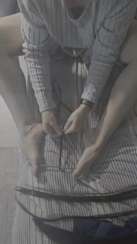 A close-up shot shows the hands of one person tying a knot with a string while another person sits on top of them, their legs straddling the person tying the knot. The person on top is wearing a grey striped long-sleeved shirt. The person underneath has their feet visible, one with a watch on the wrist. They are on a bed with striped bedding. Long dark hair is visible at the bottom of the frame.