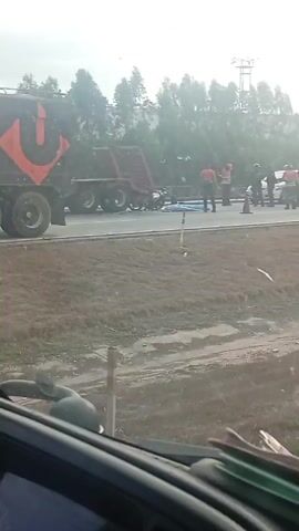 The video shows a traffic accident scene on a highway. A flatbed trailer, angled upwards, sits on the shoulder of the road. A motorcycle is visible near the trailer. A body, covered by a blue tarp, lies on the road next to the motorcycle. Two people in orange vests stand nearby, possibly first responders or traffic police. A vehicle passes in the opposite direction. The video is recorded from inside a vehicle, with the steering wheel visible. Another motorcyclist wearing a green vest and a helmet is also present at the scene.