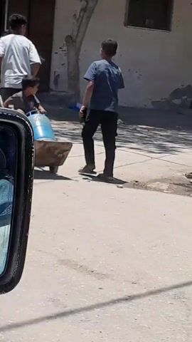 The video shows a street scene with several people. In the foreground, a child is sitting on a trolley carrying a large blue barrel. Another child walks beside the trolley. Behind them, a group of people are standing near a building entrance, some of whom appear to be observing the children. One person is smoking. There are boxes stacked near the entrance. The environment appears to be an outdoor public space, possibly a street or sidewalk in front of a shop. The lighting suggests it is daytime. The camera appears to be mounted inside a vehicle, as indicated by the reflection in the side mirror on the left.