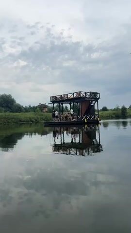 A camera pans across a body of calm water, reflecting the cloudy sky and a floating wooden structure. The structure is a multi-level raft with a dark wooden deck and railings. On the lower deck, several people are gathered around what appears to be a table, with some standing and others seated. The upper deck has railings and a small enclosed section at the back. The raft is positioned in the middle of the water, with a grassy bank and trees visible in the background, along with some distant houses. The sky is overcast with scattered clouds, and the water is still, creating a clear reflection of the raft and the sky. Two birds fly across the scene in the distance.