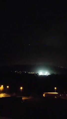 The video shows a night view of a town from an elevated perspective. In the foreground, there are a few streetlights illuminating a dark, undefined area. Beyond this, the town unfolds with numerous lights outlining streets and buildings. A brighter cluster of lights is visible in the distance, potentially indicating a larger establishment or a concentrated urban center. The sky above is dark, with a few faint lights possibly indicating distant objects or stars. The scene is still, with no discernible movement or people visible.
