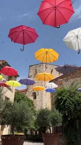 The video captures a street scene adorned with numerous colorful umbrellas suspended overhead. In the foreground, a person wearing a white t-shirt and blue shorts walks away from the camera, pushing a bicycle. The street is paved with light-colored tiles, and decorative metal fences with intricate patterns line the path. The background features an old stone building with a red brick facade, a large green wooden door, and a mural of a horse painted on the wall. Lush green foliage cascades down the side of the building, partially obscuring the door. The sky is bright blue with scattered clouds.