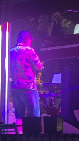 The video shows a person from behind, standing on a stage. The person is wearing a colorful, patterned short-sleeved shirt, blue shorts, and a checkered baseball cap. They appear to be performing or DJing, with some electronic equipment visible to their right. The stage is lit with vibrant purple and pink lights, and there are vertical, illuminated tubes on the left side of the frame. In the background, palm trees and a dark sky suggest an outdoor nighttime setting, possibly a club or event venue. The overall atmosphere is energetic and festive, indicated by the lighting and the presumed performance. There is no visible text in the video.
