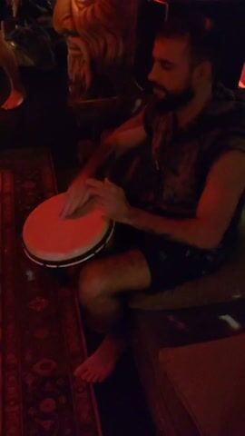 A man is seated on a dark-cushioned seat, rhythmically playing a small drum held between his knees. He is barefoot and wearing dark shorts and a sleeveless shirt with a camouflage pattern. He has a beard and his face is visible in profile as he focuses on the drum.  The environment is dimly lit with a red light illuminating the area where he is sitting, and patterned rug is visible on the floor beneath him. Other people, also barefoot, are visible in the background, moving slightly. Behind the man, there appears to be a carved, or decorated piece of furniture. The focus remains on the man and his drumming.