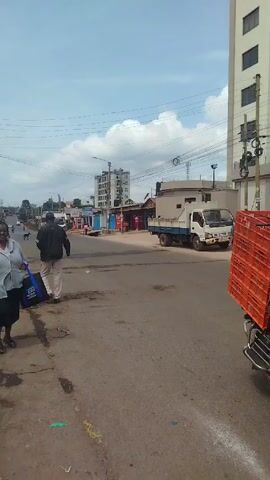 A medium-angle shot shows a street scene on a sunny day. The road is paved but appears worn and uneven. On the left sidewalk, a person is walking, carrying a dark blue tote bag with white text. Another person, partially cropped, is standing near them.

Across the street, a blue, white, and orange minibus with "PETANNS" written on its side drives past from left to right. Further down the street, a small white truck is parked. A man is walking toward the truck.

In the background, there are various buildings, including a tall, light-colored building on the right with visible windows and utility poles. On the left side of the street, there are smaller buildings and structures, including a single-story building with red and blue coloring. The sky is blue with some white clouds. Utility wires stretch across the scene.