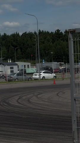 A red BMW M3 is drifting in a parking lot. The car is kicking up smoke from its tires as it slides around a corner. The car has several stickers on its windshield and sides, including one that says "Valvoline" and another that says "Westmotors." The parking lot appears to be an empty industrial area with a fence and trees in the background. There are other cars parked in the background, as well as a few people standing around. The sky is blue with some clouds.