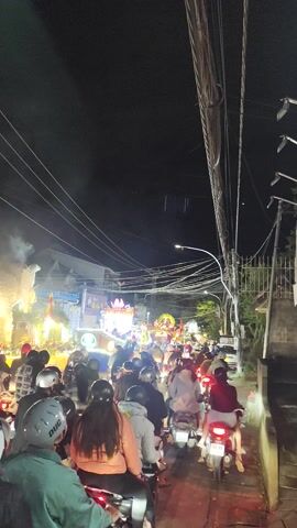 This vertical video captures a lively night scene on a bustling street, indicative of a festive event or parade in Lang Biang - Đà Lạt, Vietnam.

The frame is filled with a dense crowd of people, predominantly on motorbikes, packed closely together on what appears to be a street or road. Many individuals are wearing helmets of various colors, including black, blue, and white. The motorbikes appear to be moving slowly or are stationary, suggesting heavy traffic or participation in a procession.

In the center of the activity, a brightly lit and elaborately decorated float or display is the focal point. It features numerous lights and what appear to be floral or decorative arrangements, emitting a significant amount of white smoke or fog that rises into the night air. This smoke suggests the use of pyrotechnics, fog machines, or incense, contributing to a celebratory atmosphere.

Overhead, a complex web of power lines stretches across the upper left portion of the frame, contrasting with the dark night sky. Buildings with illuminated signage line the street in the background. Some Vietnamese text is partially visible on a sign to the left of the main display, though not entirely legible.

The overall impression is one of a vibrant, crowded, and festive gathering under the cover of night, illuminated by artificial lights from street lamps, motorbikes, and the central display.