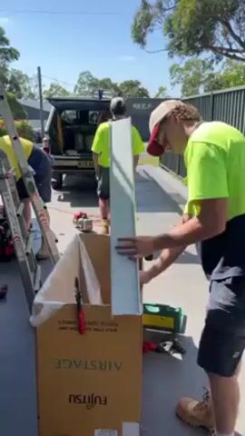 The video shows three men working outdoors on a sunny day. One man, wearing a lime green t-shirt, dark shorts, and a baseball cap, stands in the foreground. He is holding a long, gray, rectangular piece of metal and appears to be aligning it with the edge of a large cardboard box labeled "AIRSTAGE" and "FUJITSU". A second man, also in a lime green t-shirt, is working on a ladder in the background to the left. A third man, wearing similar attire, is visible working by a white truck parked further back. The ground is paved, and various tools are scattered around, including a drill, pliers, and a green toolbox. A green fence runs along the background, and trees are visible in the distance under a clear blue sky. The scene gives the impression of a construction or installation project in progress.