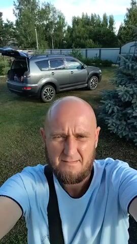 A bald, bearded man wearing a light blue t-shirt and a black strap across his chest is taking a selfie video outdoors. Behind him, a dark gray SUV is parked with its trunk open. The setting appears to be a rural backyard with a fence, green grass, and several trees, including a large blue spruce. The man smiles and turns the camera to capture different views of his surroundings.
