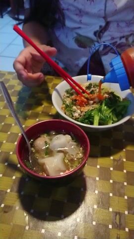 The image presents an overhead view of an indoor dining scene, likely featuring an Asian meal. A person's hand and forearm, clad in a white garment with a floral pattern, are visible on the left side of the frame. The person is actively engaging with the food, holding a pair of red chopsticks and squeezing a red condiment bottle with a blue cap over a bowl of noodles.

The main dish is served in a large, white, oval-shaped bowl. It contains a substantial portion of noodles, mixed with vibrant green leafy vegetables (resembling bok choy or similar greens), and topped with what appears to be a chili-infused sauce or minced meat mixture. The red chopsticks are positioned within this bowl, with some noodles lifted up, demonstrating the texture of the dish.

Adjacent to the noodle bowl, on the right, is a smaller, dark red or maroon bowl containing a clear broth. This soup bowl holds two prominent, white, folded dumplings or wontons, several smaller round items that look like meatballs, and is garnished with chopped green onions. A silver spoon is resting inside this soup bowl.

The meal is arranged on a table covered with a checkered tablecloth, featuring a pattern of alternating yellow and brown squares. Notable shadows, particularly from the smaller bowl and its contents, are cast upon the tablecloth, indicating a light source from the left side of the scene. The overall impression is of a casual meal setting. No discernible text is visible within the image.