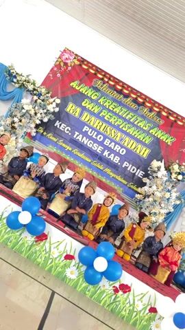 The video captures a graduation ceremony for young children at RA Darussa'adah in Pulo Baro, Tangse District, Pidie Regency, Indonesia. A group of children, dressed in traditional attire, are seated on a stage in front of a large banner that reads "Selamat dan Sukses Ajang Kreativitas Anak dan Perpisahan RA Darussa'adah Pulo Baro Kec. Tangse Kab. Pidie." Below this, smaller text states, "Do'a Kami Semoga Kesuksesan Selalu Menyertaimu Selama." The banner is adorned with floral motifs.

The stage is decorated with blue and white fabric drapes, floral arrangements, and several clusters of blue and white balloons. The children are arranged in two rows, with some wearing traditional headwear and ornate clothing. They appear to be posing for the camera, with some looking directly at it. The overall atmosphere seems celebratory and formal, typical of a school event. The lighting suggests an indoor setting, likely during the day. The children's expressions are mostly neutral or slightly posed smiles, indicating they are participating in a structured event. The presence of the banner and the children's attire strongly suggest this is an Indonesian cultural event, specifically a graduation or end-of-year ceremony for a kindergarten or early childhood education center.