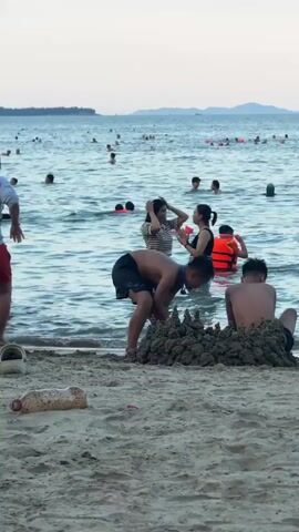 The media file depicts a crowded beach scene with numerous people in the shallow water, engaged in recreational activities. The water is calm, suggesting pleasant weather, and the lighting indicates it's likely late afternoon or early evening as the sun begins to set, casting a soft glow over the scene. The sky is visible in the background with a faint horizon line of landmasses in the distance, possibly islands or a coastline.

In the foreground, a partially constructed sandcastle is visible on the sandy shore, hinting at family fun and creative play. Several individuals are actively participating in the water. A young boy, shirtless and wearing dark shorts, is positioned near the sandcastle, appearing to be either playing or interacting with others in the water. Another boy, also shirtless, is seated in the foreground, partially obscured by the sandcastle.

Among the crowd in the water, several people are wearing bright orange life vests, suggesting caution or the presence of designated swimming areas. A woman in a striped shirt and dark skirt is standing in the water, with another person behind her. To the left, a person wearing a grey shirt and red shorts is partially visible, also standing in the water. The overall atmosphere appears lively and relaxed, with people enjoying a day at the beach. There is no visible text in the media file. The location context provided indicates the scene is likely Bac Nha Trang Ward, Vietnam, known for its beaches.