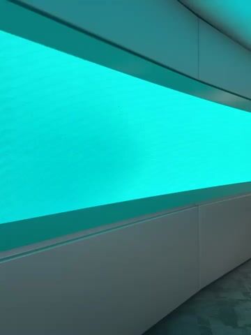 This is a short video clip that appears to be filmed indoors, likely in a modern setting with light-colored wall panels. The primary subject is a large, rectangular digital screen displaying a vibrant turquoise background. Initially, the screen is a solid color, but at around the 00:03 mark, a pattern of small, colorful, rectangular objects appears. These objects are arranged in neat rows and columns across the screen, resembling a collection of pills, candies, or perhaps small stickers. They vary in color, including red, yellow, blue, and white. The lighting is artificial and bright, casting a cool, even glow. There is no indication of the time of day or weather. No people are visible in the clip. The setting appears to be clean and contemporary, with some textured flooring visible at the bottom right of the frame. No text is present on the screen or in the surrounding environment.