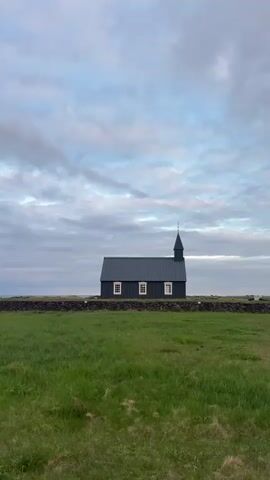 A vertical shot depicts the iconic black church of Búðir (Búðakirkja) in Iceland, standing prominently in a vast green field. The church, painted a deep, dark hue, features a steep roof and a small steeple with a cross at its peak. Several rectangular white-framed windows are visible on its side, providing a stark contrast to the dark exterior.

A low, rustic stone wall runs horizontally across the mid-ground, separating the foreground's bright green grass from the church and the distant horizon. The grass in the foreground appears lush and untamed.

The sky above is overcast with soft, diffused light, showing a mix of grey and lighter cloud formations, with hints of blue peaking through. The overall atmosphere suggests a quiet, somewhat serene moment, possibly in the early morning or late afternoon given the soft illumination. There are no people visible in the scene. The landscape conveys a sense of peaceful solitude typical of the Icelandic countryside.