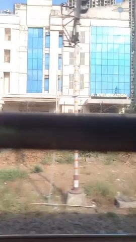 The video shows a view from a moving train, looking out at the landscape. Initially, the camera focuses on a multi-story building with blue windows, identified as "TOYO TECHNOLOGY," followed by glimpses of other buildings, some under construction, featuring cranes. The vegetation is visible in the foreground, and utility poles appear throughout the shot. The sky is clear and blue. The camera passes structures quickly, offering brief views of the urban landscape.