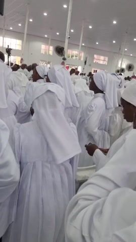 The video depicts a large gathering of people, predominantly adults, within a spacious, brightly lit indoor hall. All individuals are uniformly dressed in white robes or gowns, with many also wearing white head coverings. They are standing and facing forward, actively participating in what appears to be a religious service or worship event, as indicated by the harmonious singing audible in the background.

The setting is a large hall with white walls, a white ceiling equipped with recessed lighting and ceiling fans, and high windows providing natural illumination. In the background, a camera or video equipment is mounted on a pole, and a monitor screen is visible on a pillar, suggesting the event might be recorded or broadcast. Some attendees are seen holding mobile phones. The overall atmosphere conveys a sense of collective devotion and reverence. Given the context of Anantigha, Nigeria, and the distinctive white attire, this gathering is likely a church service, possibly of a denomination known for such garments. No visible text is present in the frame.