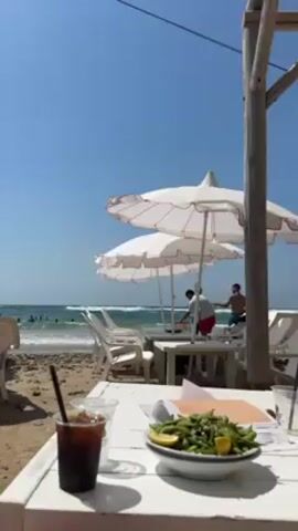 The video shows a beach scene, with a table in the foreground set with a drink and a bowl of edamame. In the background is the ocean, beachgoers, and two white umbrellas covering outdoor seating. The sky is clear and blue. Two people are seen standing at another table. A wooden post is visible on the right side of the shot.