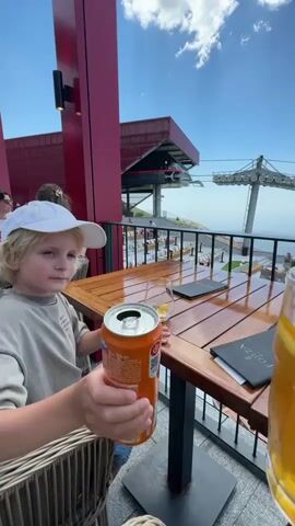 This media file shows a young boy with blonde hair wearing a white baseball cap and a beige t-shirt, sitting at a wooden table outdoors. He is holding an orange can of Fanta in his right hand and a glass of a yellow beverage with lemon slices and ice in his left. His gaze is directed towards the viewer, and he appears to be smiling faintly. In the background, another person's hand is visible holding a glass, suggesting a toast.

The setting is a restaurant or cafe with outdoor seating, overlooking a scenic view. The architecture of the building is modern, with a prominent red facade and a structure that resembles a ski lift station or cable car terminal. The weather appears to be sunny and pleasant, with a blue sky and scattered clouds. The time of day seems to be daytime, likely afternoon, given the lighting.

Visible text includes "Fanta" on the can and "FORZA" on a menu on the table. The location is identified as Kuk, Montenegro. The overall mood is one of enjoyment and leisure, possibly a family outing or vacation.